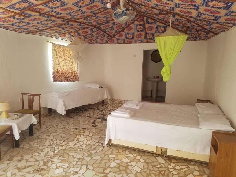 Una habitación con cama, una mesa y una silla. Hay una ventana con cortinas y un ventilador en el techo.