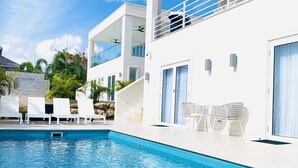 Un espace moderne aménagé au bord de la piscine, avec une piscine à l’eau d’un bleu limpide, des chaises longues blanches et un bâtiment à deux étages doté de balcons.