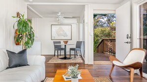 A living room with a white sofa, a coffee table with a plant, a dining area with a table and chairs, and a view of a garden through an open door.