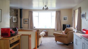 A cozy living room with a sofa, armchair, and a small table with a red appliance. A window with curtains and a lamp above it is visible. - The Coach House - UK30813 (Thornthwaite, near Keswick)