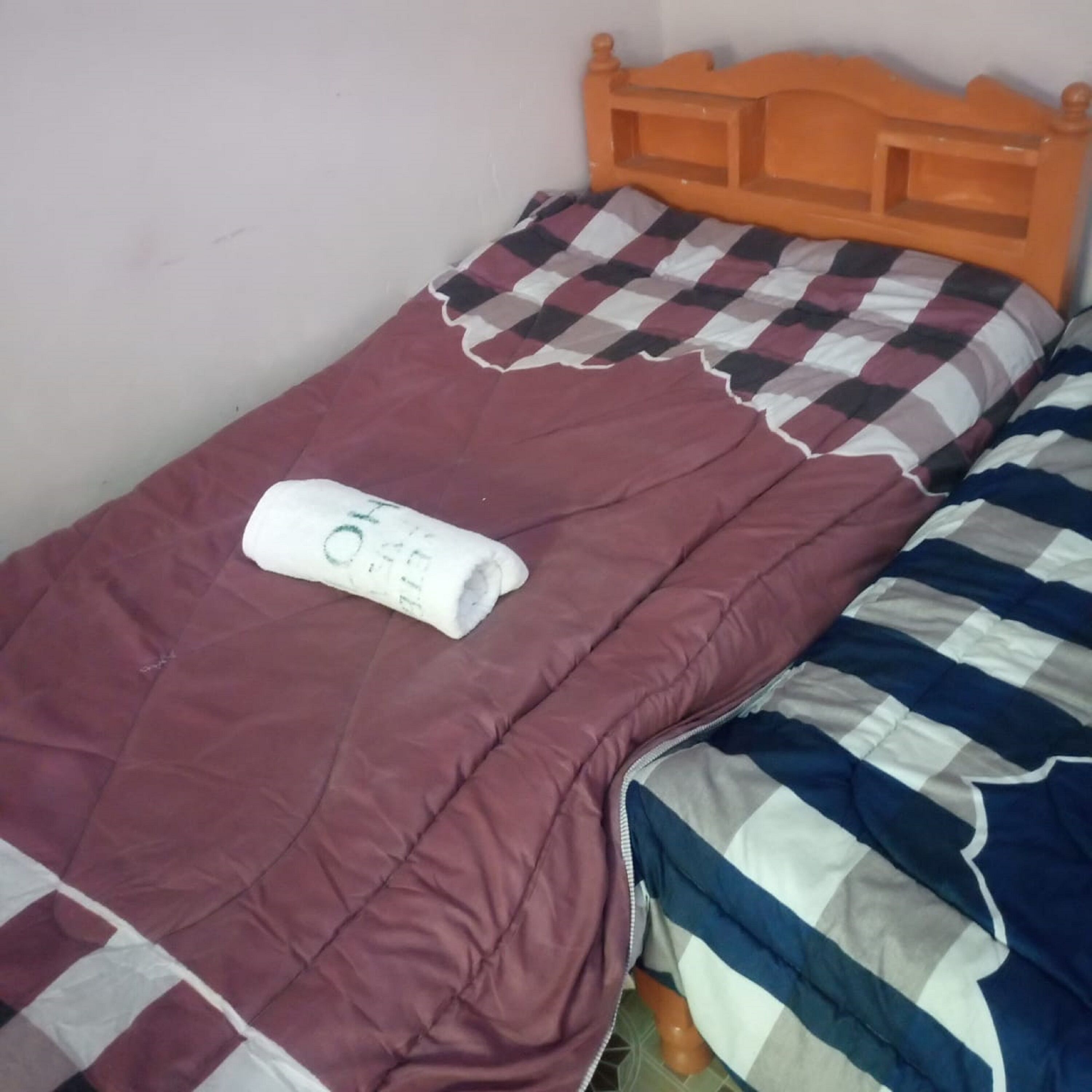 A single bed with a checkered pillow and a rolled-up towel on a maroon bedspread.