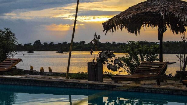 A poolside area with a thatched umbrella, lounge chairs, and a view of a body of water during sunset.