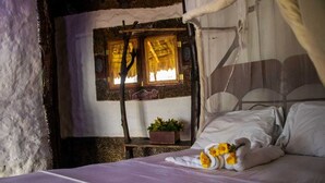 A rustic bedroom with a bed, a window with a wooden frame, and a potted plant on a small table beside the bed.