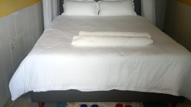 A neatly made bed with a white bedspread and pillows, a pair of blue and red slippers on a checkered rug, and a window with white curtains. - Venus lodge Kikopeye (Gilgil)