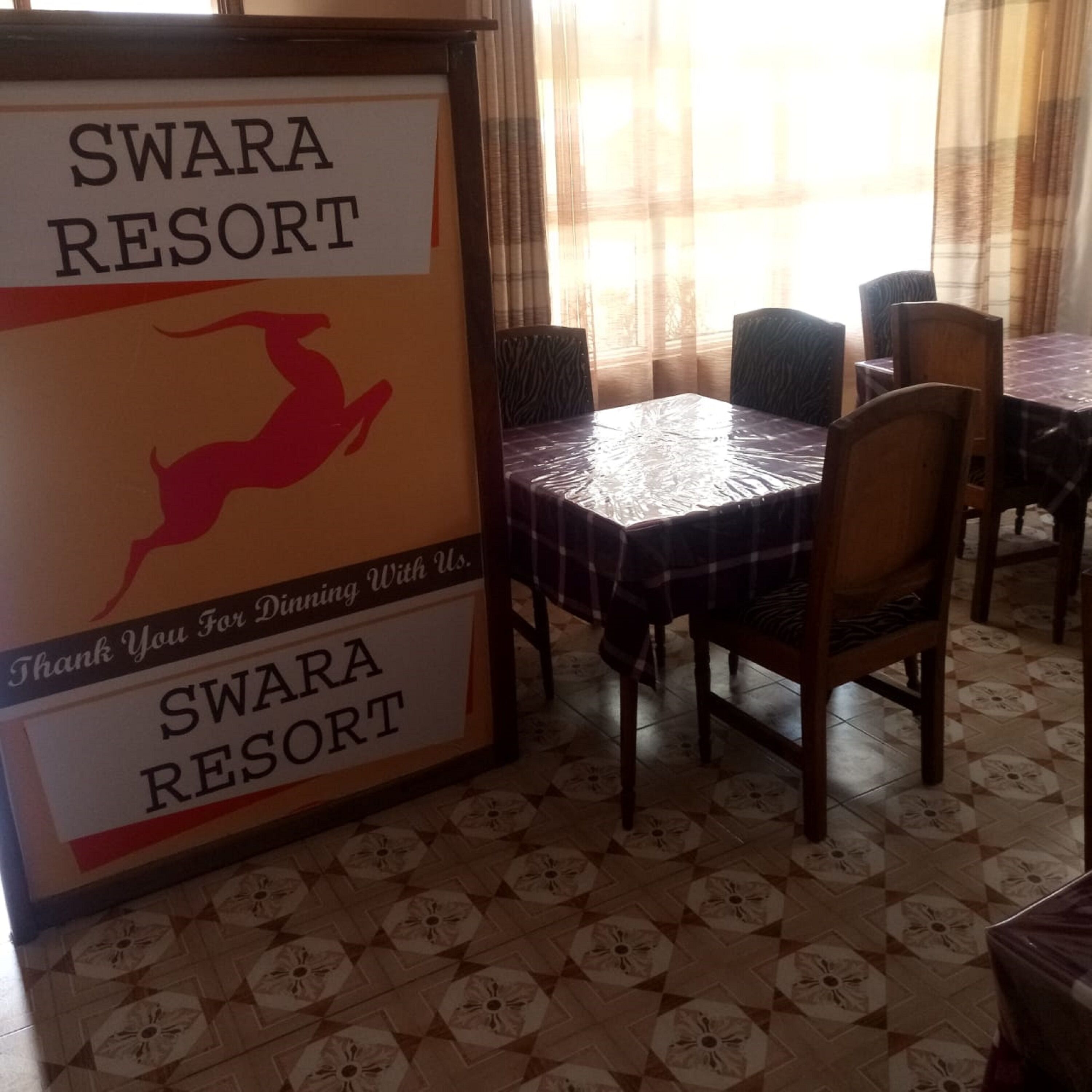 A dining area with a sign reading "SWARA RESORT" and a thank you message. Tables are covered with checkered tablecloths, and chairs are arranged around them.