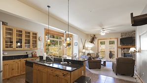 A spacious kitchen with wooden cabinets, a large island, and a dining area with a fireplace. There is a ceiling fan and a television mounted on the wall. - Beautiful mountain views from this immaculate 3 bedroom ski and summer condo (Whitefish)