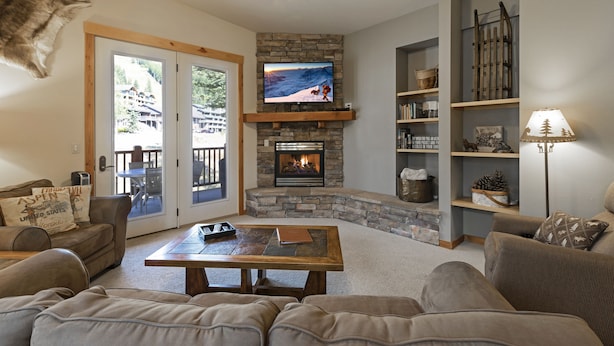 A cozy living room with a stone fireplace, a television mounted above it, and a bookshelf filled with books and decorative items. - Beautiful mountain views from this immaculate 3 bedroom ski and summer condo (Whitefish)
