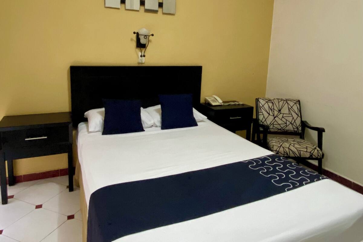 Una habitación de hotel con una cama grande, una mesita de noche, una silla y un teléfono fijo en la pared.