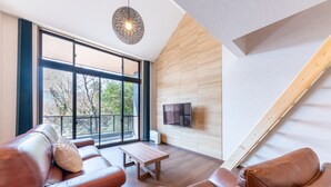 Una sala de estar moderna con sofás de cuero, una mesa de centro de madera, un televisor grande y vistas a árboles a través de amplias ventanas.