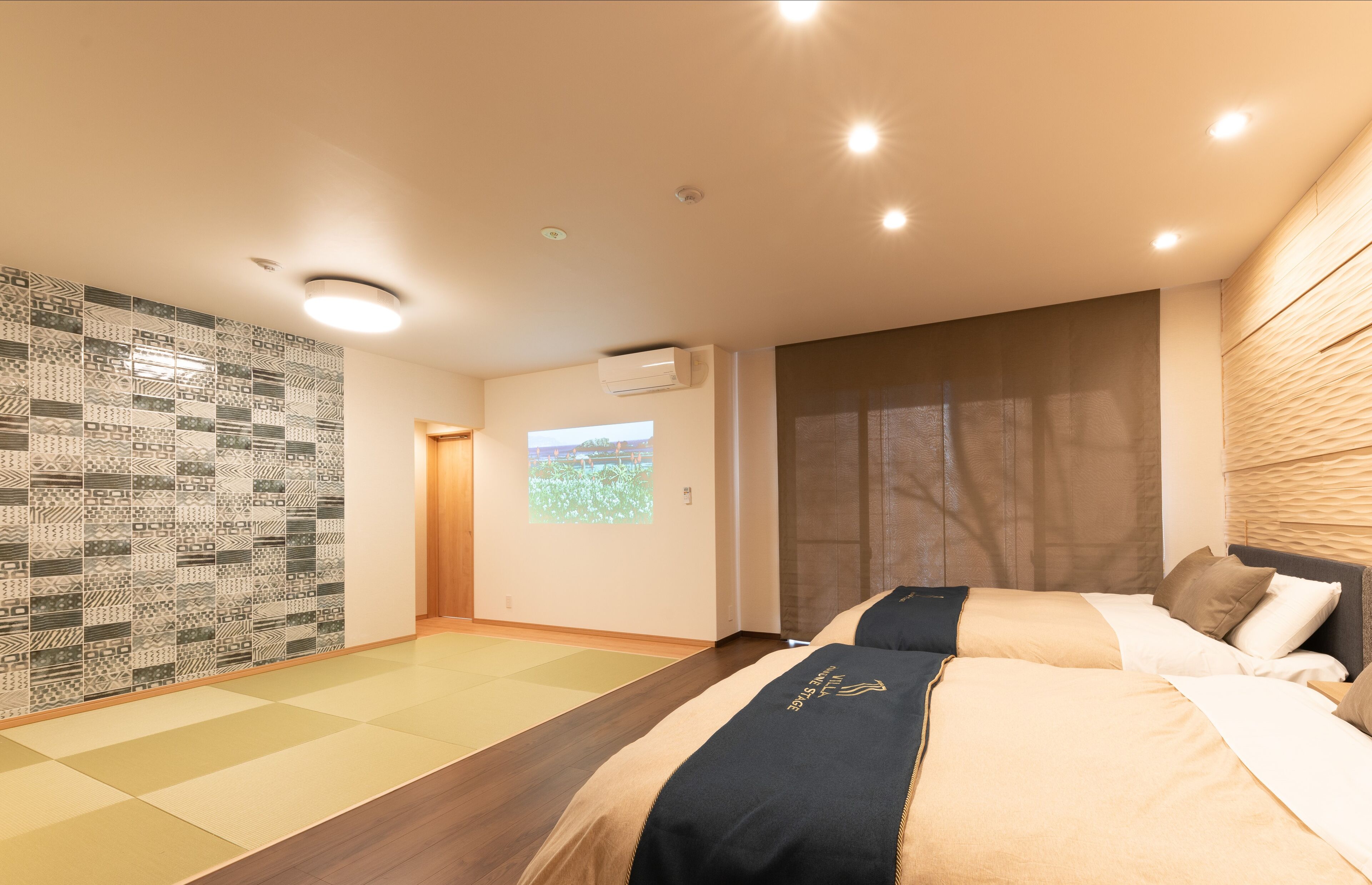 A hotel room with two beds, a large window, a projection screen, and a patterned wall.