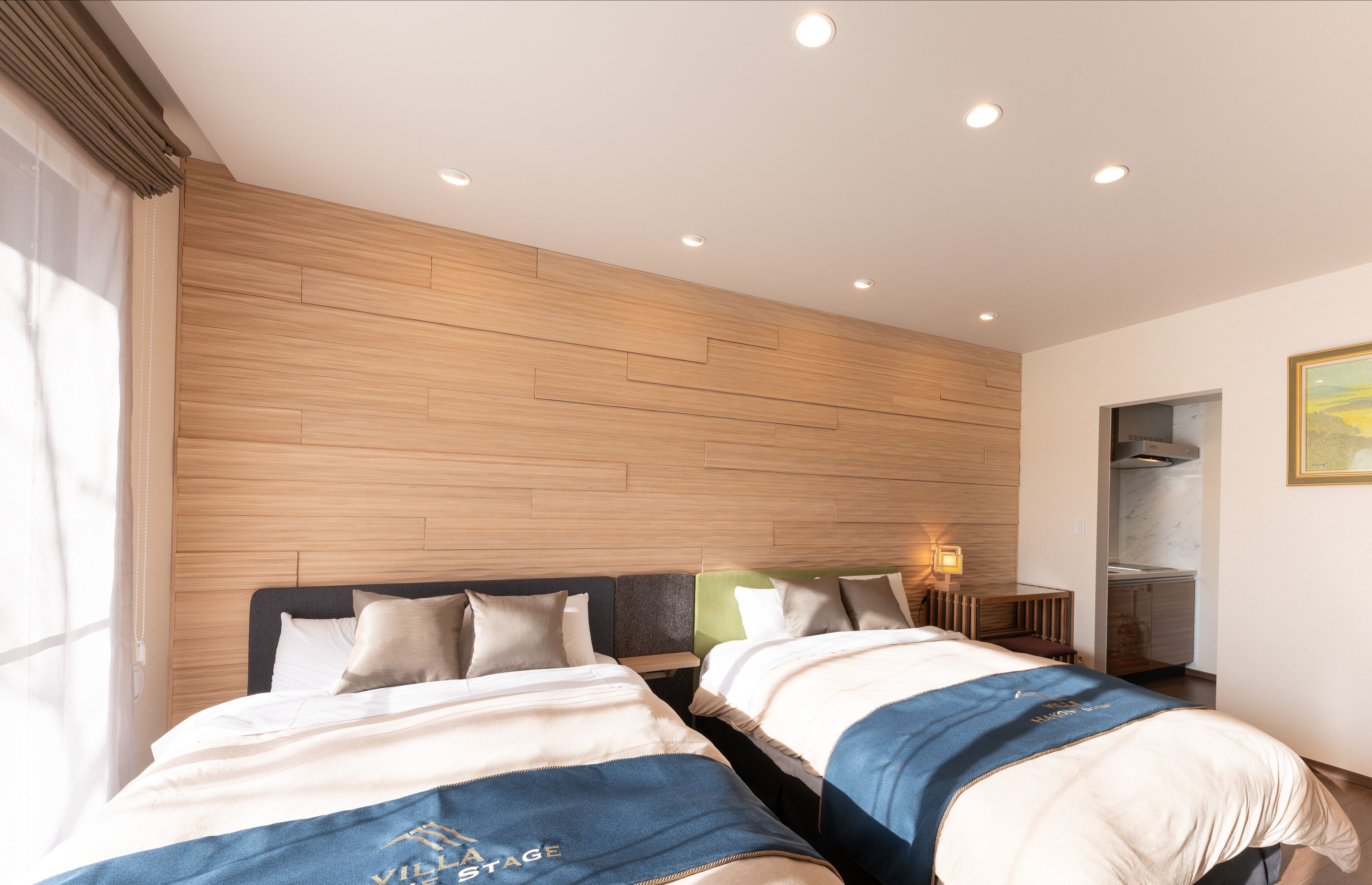 Habitación de hotel con dos camas, cabecera de madera, mesita de noche con lámpara y ventana con cortinas.