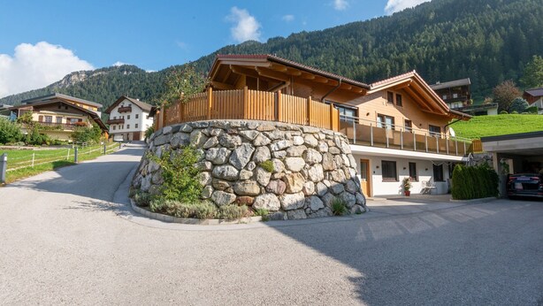 A mountain lodge with a stone wall entrance, wooden balconies, and a view of the surrounding forested mountains.