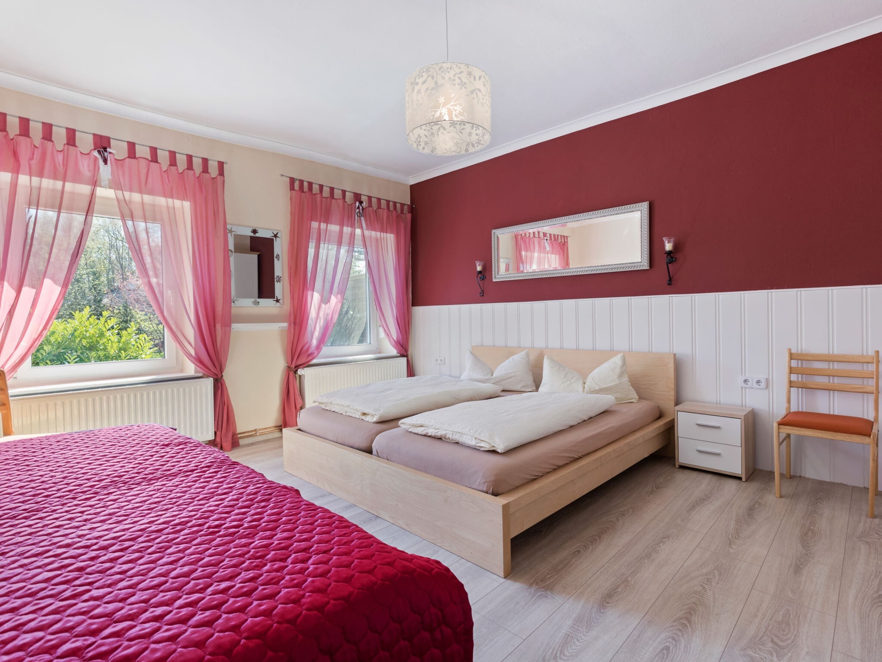 Une chambre à coucher comprenant un lit, des tables de chevet, une chaise, une fenêtre avec des rideaux, un miroir et un luminaire suspendu.