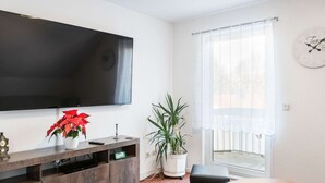 Un salon équipé d’un téléviseur à écran plat, d’une table basse, d’un canapé, d’une plante en pot et d’une fenêtre avec des stores.