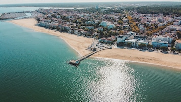 Vista aérea de un pueblo costero con un muelle que se adentra en el mar, rodeado de edificios residenciales y una playa.