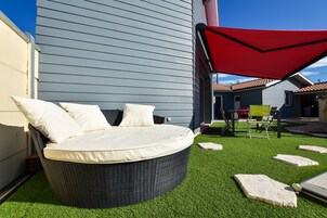 A round wicker bed with white cushions, a red canopy, and artificial grass in a courtyard.