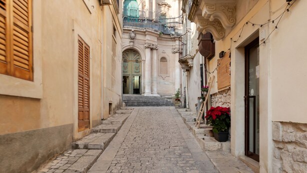 A narrow cobblestone street flanked by historic buildings with ornate facades and a central staircase leading to an upper level.