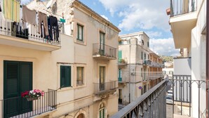 A narrow street with buildings featuring balconies, green shutters, and a pedestrian walkway.