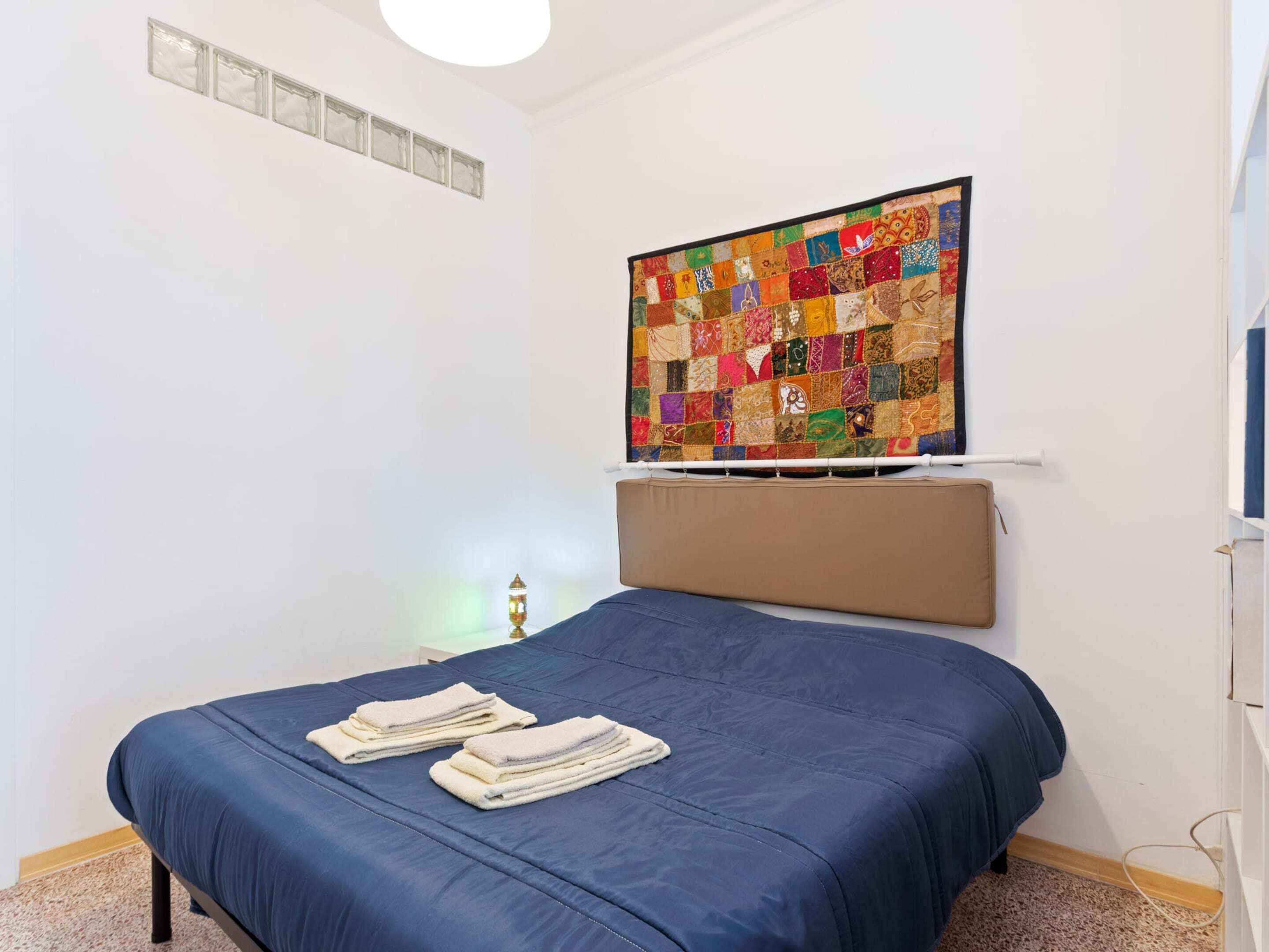 A bedroom with a blue bedspread, a quilt hanging on the wall, and a bedside table with a lamp.