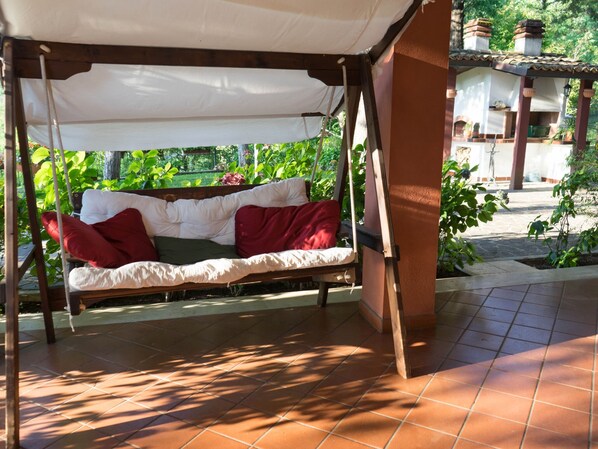 A covered outdoor seating area with a swing-style sofa, a tiled floor, and a garden view. - Villa in Tagliacozzo With Pool and Nature (Tagliacozzo)