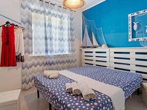 A bedroom with a bed, a bench, a window with curtains, a wall with a sailboat decoration, and a rack with clothes.