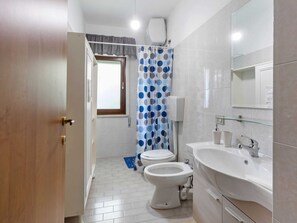 A bathroom with a toilet, bidet, and sink. There is a window with a curtain, and a mirror above the sink.