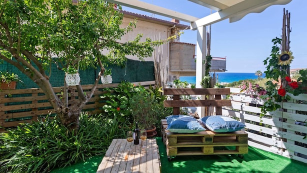 A wooden deck with a green artificial turf, a wooden table with wine glasses, and a bench with pillows. - Cosy Holiday Home in Sanremo With Sea Beach Nearby (Alcamo)