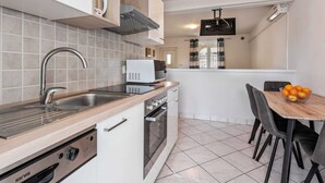 A modern kitchen with a stainless steel sink, a built-in oven, and a microwave. There is a dining area with a wooden table and chairs, and a window with curtains.