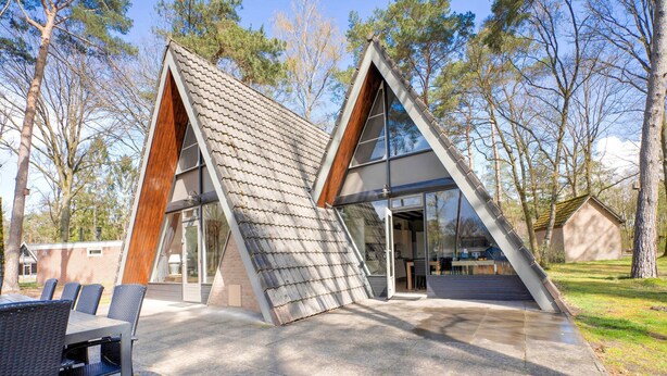 A modern, triangular house with a sloped roof, large windows, and a patio area with outdoor furniture.