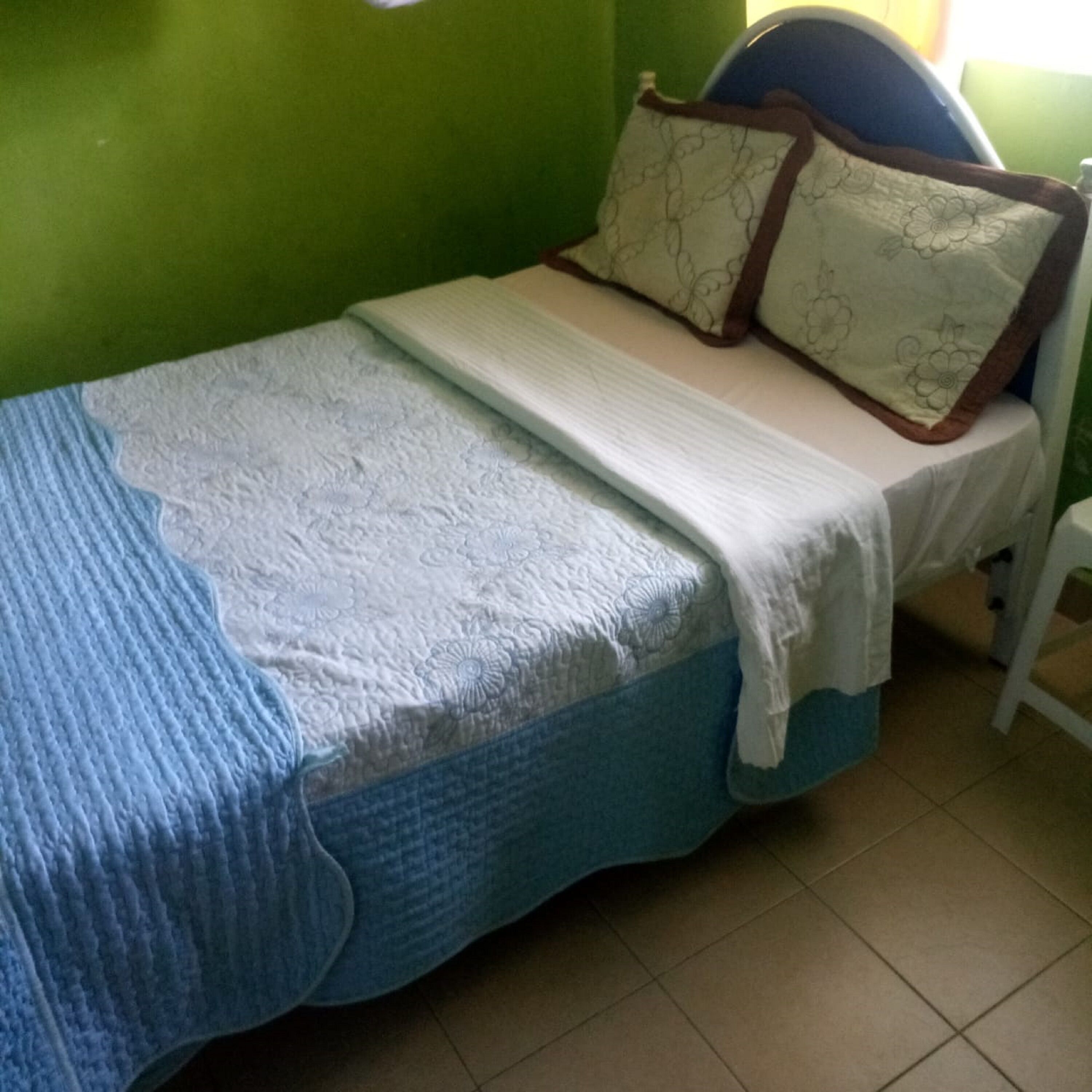 Una cama individual con una manta azul y una colcha blanca, un almohadón con estampado floral y un cabecero con detalles marrones.