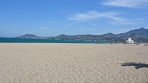 Una playa con un edificio blanco, montañas al fondo y cielo azul despejado.