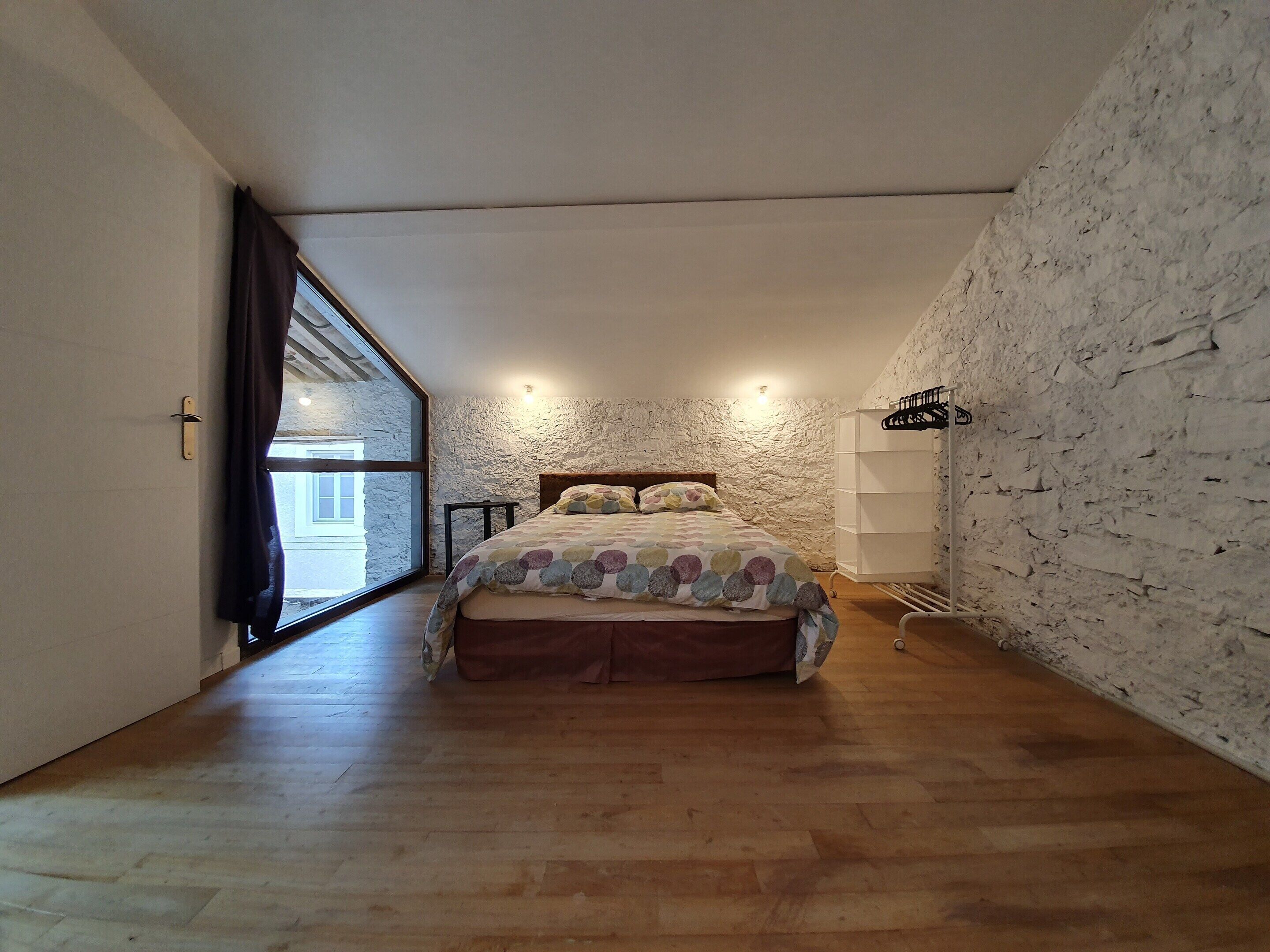 A bedroom with a bed, a window, a wall-mounted shelf, and a wooden floor.