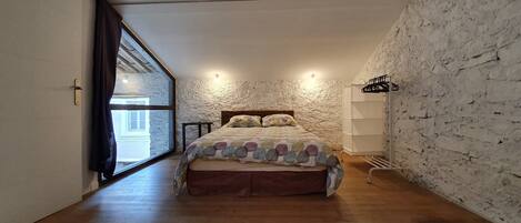 A bedroom with a bed, a window, a wall-mounted shelf, and a wooden floor.