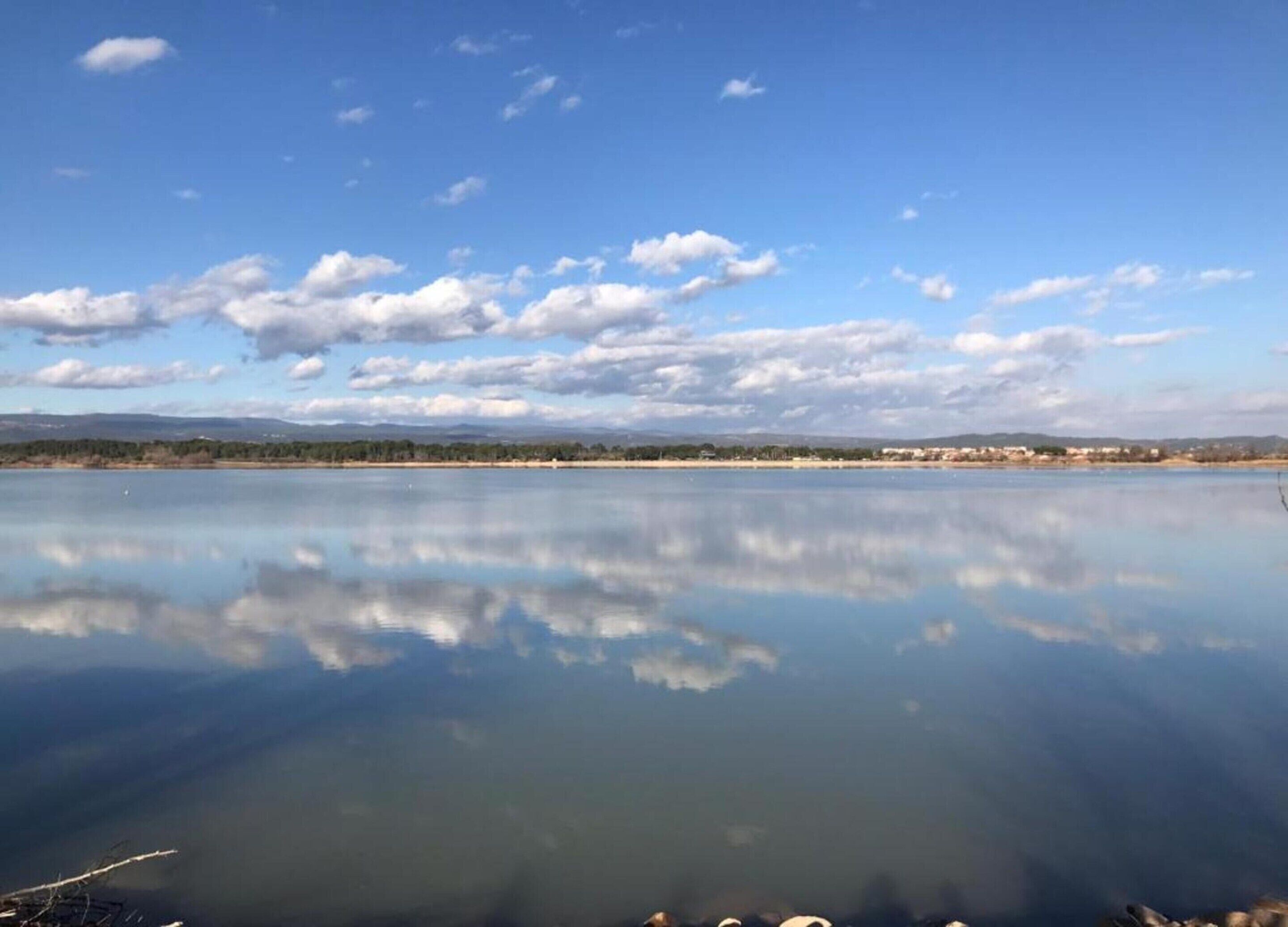 A calm lake with a clear reflection of the sky and clouds, surrounded by a flat landscape with distant buildings.