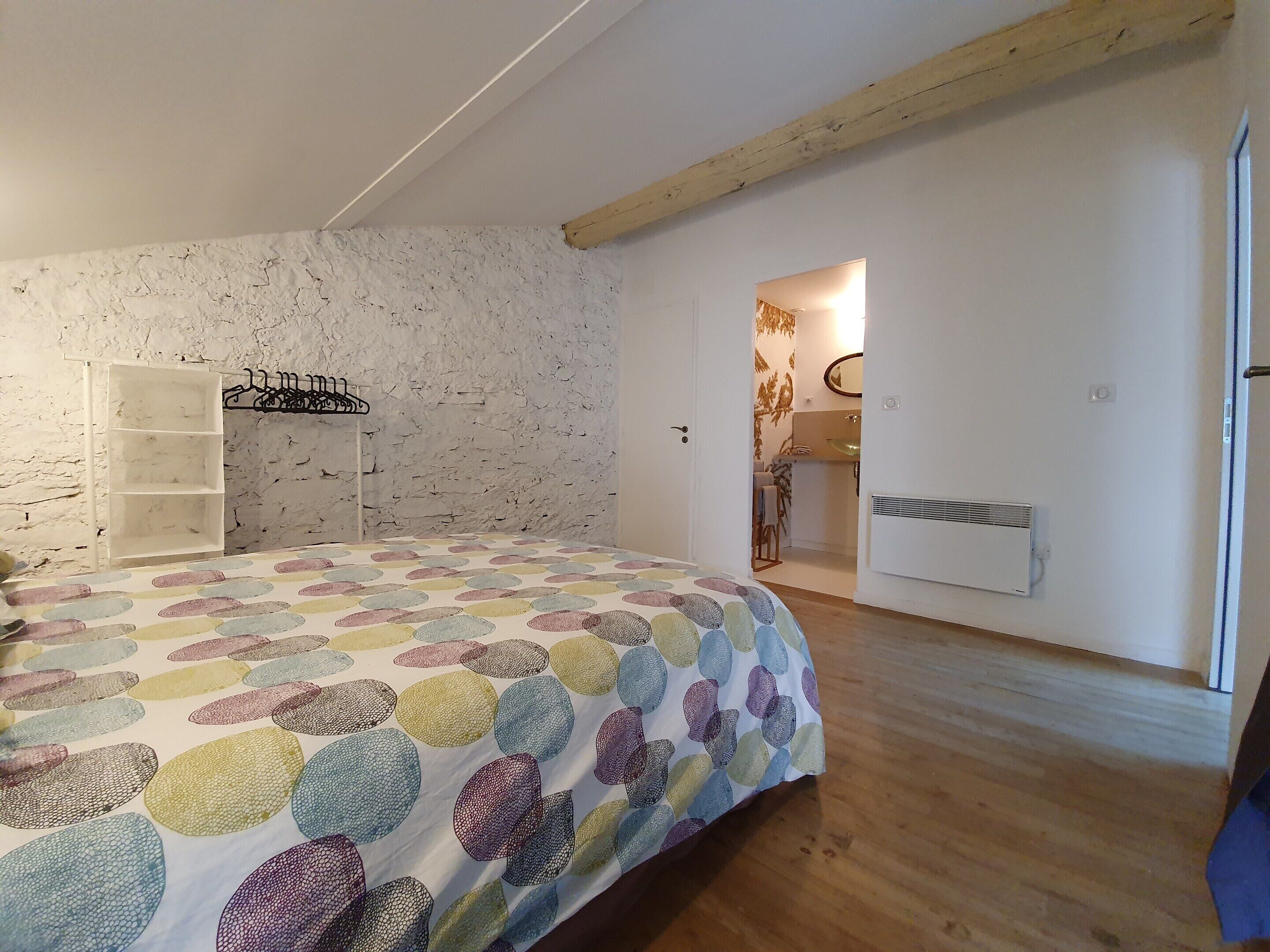 A bedroom with a bed, a white wardrobe, a wooden ceiling beam, and a bathroom visible through an open door.