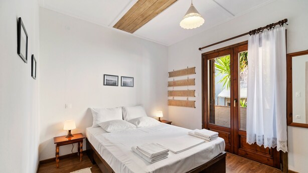 A bedroom with a wooden bed, white bedding, a nightstand with a lamp, a wooden floor, and a view through a window with curtains. - Snug Apartment in Agios Nikolaos With Garden (Sithonia)