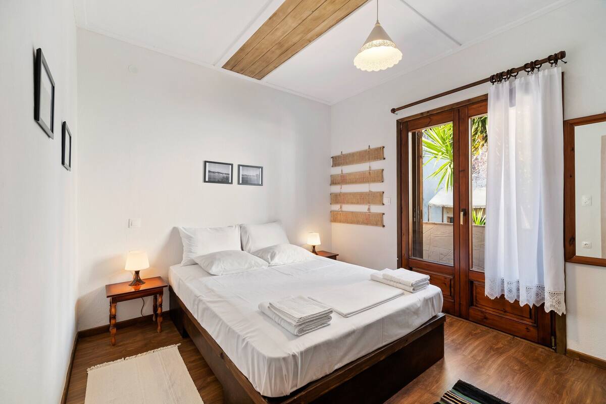 A bedroom with a wooden bed, white bedding, a nightstand with a lamp, a wooden floor, and a view through a window with curtains.