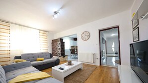 Una sala de estar moderna con un sofá gris, una mesa de centro blanca y un televisor. Hay un armario de madera y un reloj en la pared.
