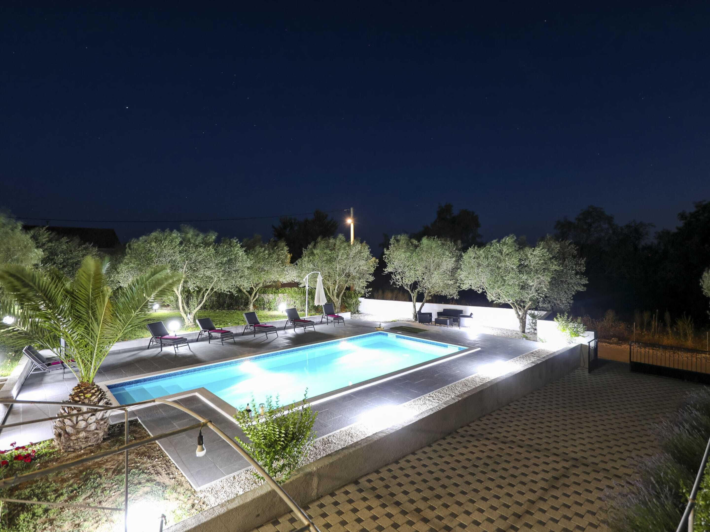 A swimming pool with lounge chairs and umbrellas, surrounded by trees and a night sky.
