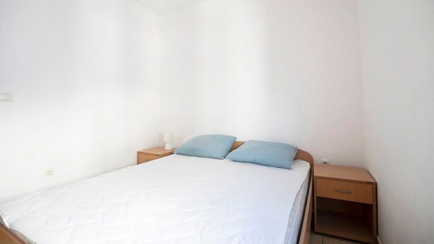 A single bed with a white mattress and two pillows, a wooden nightstand with a lamp, and a small drawer unit. - Tasteful Apartment in Zadar With Balcony (Zadar)