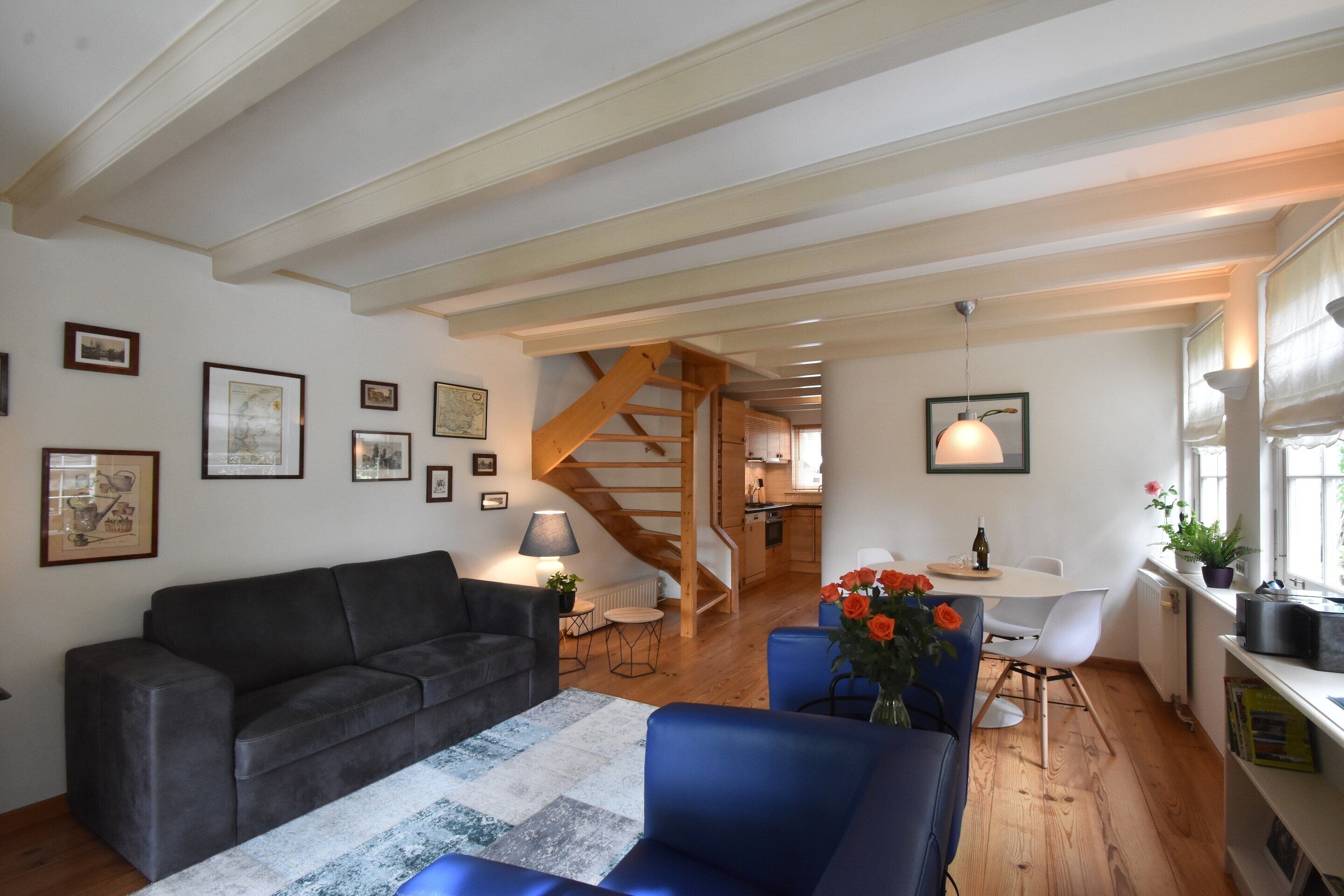 A cozy living room with a sofa, a blue armchair, and a coffee table. There is a staircase leading to an upper level, and a dining area with a table and chairs.