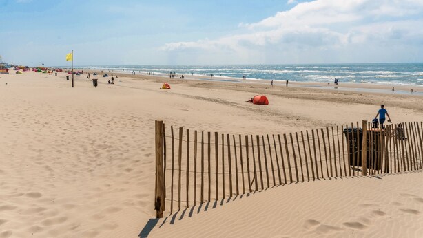 Une plage avec une clôture en bois, un drapeau jaune et des personnes qui se promènent le long du rivage.