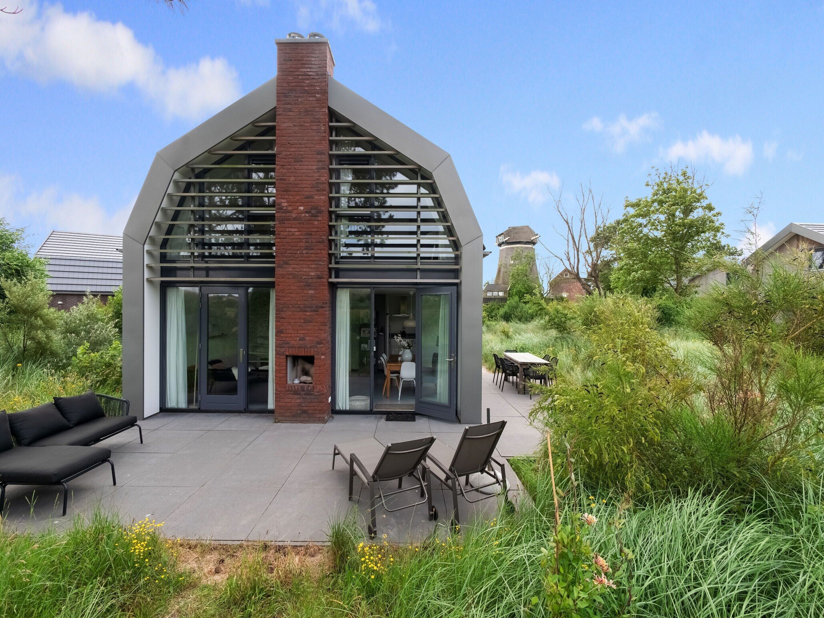 Ein modernes Haus mit einem Ziegelkamin, großen Fenstern und einer Terrasse mit Gartenmöbeln.