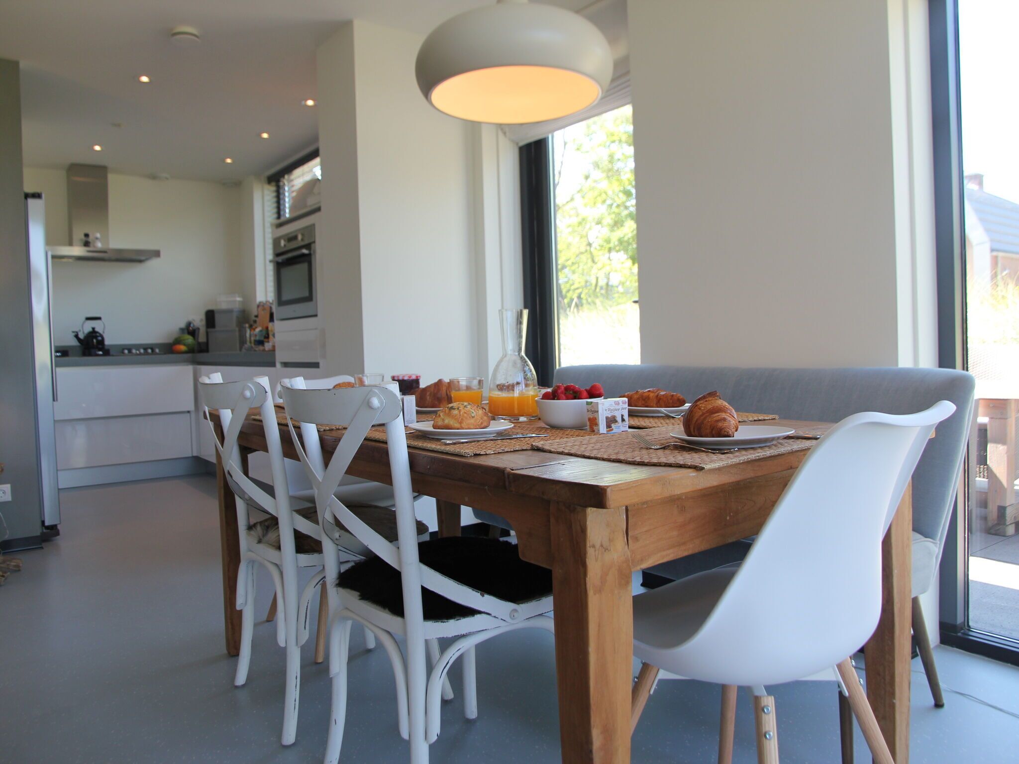 A modern dining area with a wooden table, white chairs, and a breakfast setting including croissants, fruit, and juice.