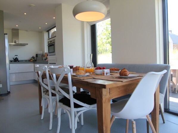 A modern dining area with a wooden table, white chairs, and a breakfast setting including croissants, fruit, and juice. - Dune Villa in Egmond aan Zee (Egmond aan den Hoef)
