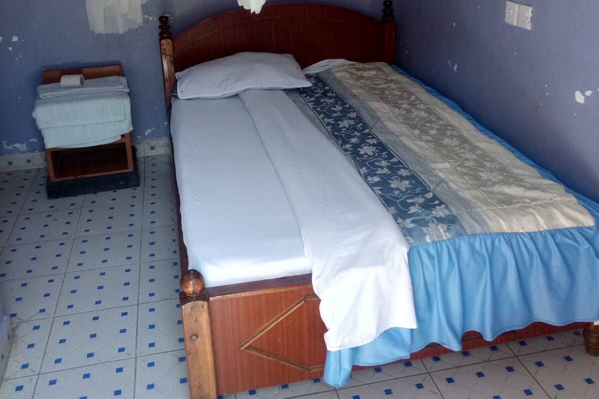 A single bed with a wooden frame, a blue and white bedspread, and a white pillow. There is a small wooden chest of drawers to the left of the bed.