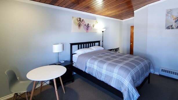 Un dormitorio con techo de madera, una cama con colcha escocesa, una mesita de noche con lámpara, una silla y una pequeña mesa redonda.