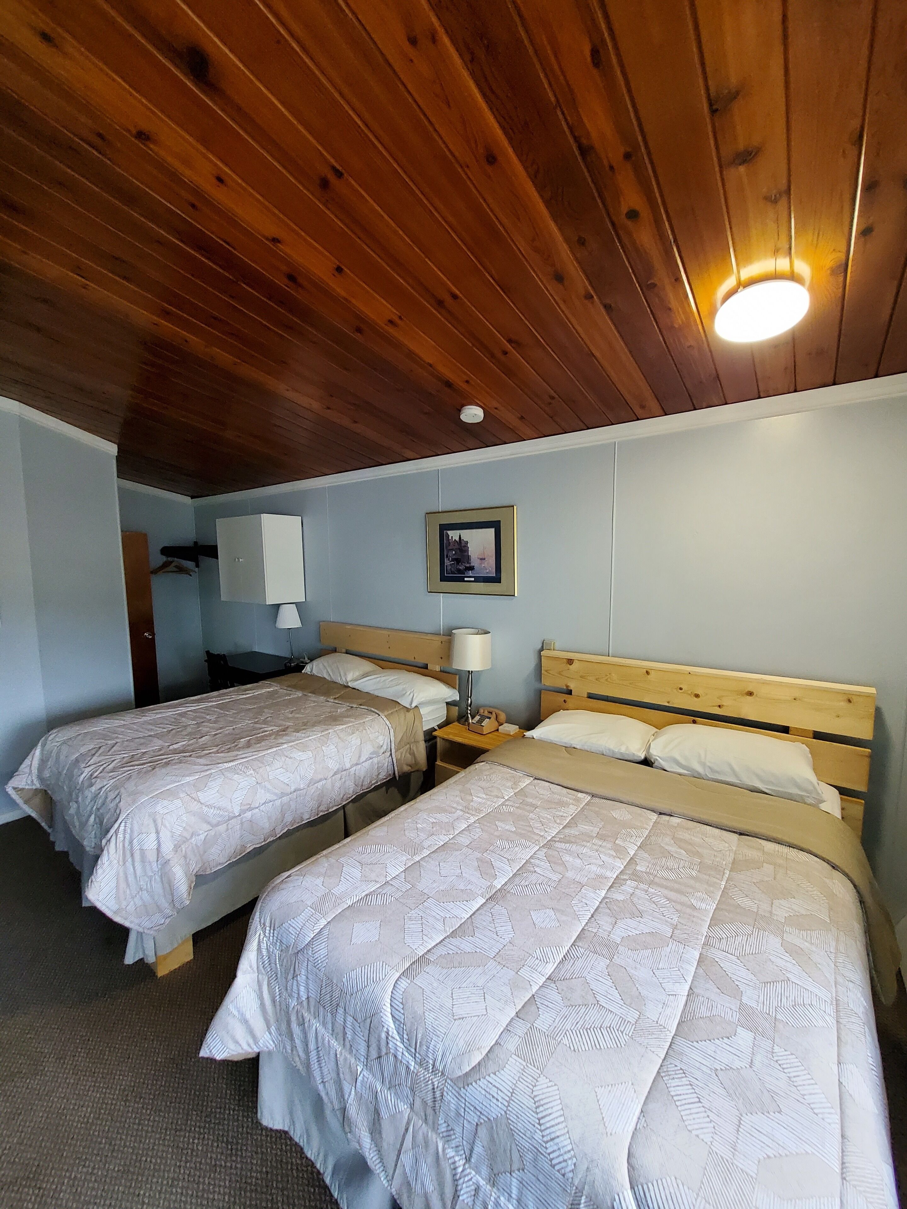 A room with two beds, wooden ceiling, a framed picture on the wall, and a nightstand with a lamp.