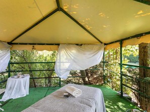 A tented outdoor area with a bed, a table with items, and a view of greenery.