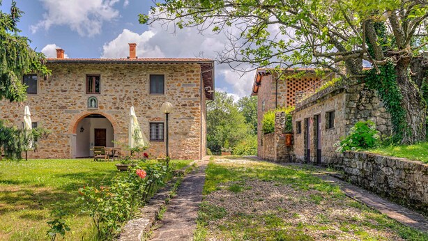 Un bâtiment en pierre avec une entrée centrale, un petit patio doté d’une table et de chaises, et un chemin menant à un jardin.